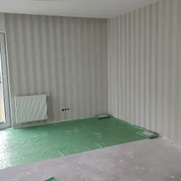 Pokój w trakcie przygotowania do malowania lub tapetowania: ściany w pionowe pasy, podłoga zabezpieczona folią, widoczne puszki z farbą lub klejem.