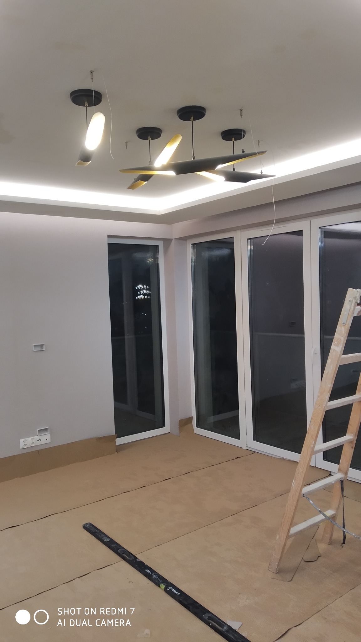 Wnętrze pokoju w trakcie prac wykończeniowych: podłoga zabezpieczona kartonem, drabina oparta o ścianę, poziomica leżąca na podłodze, sufit z nowoczesnym oświetleniem.