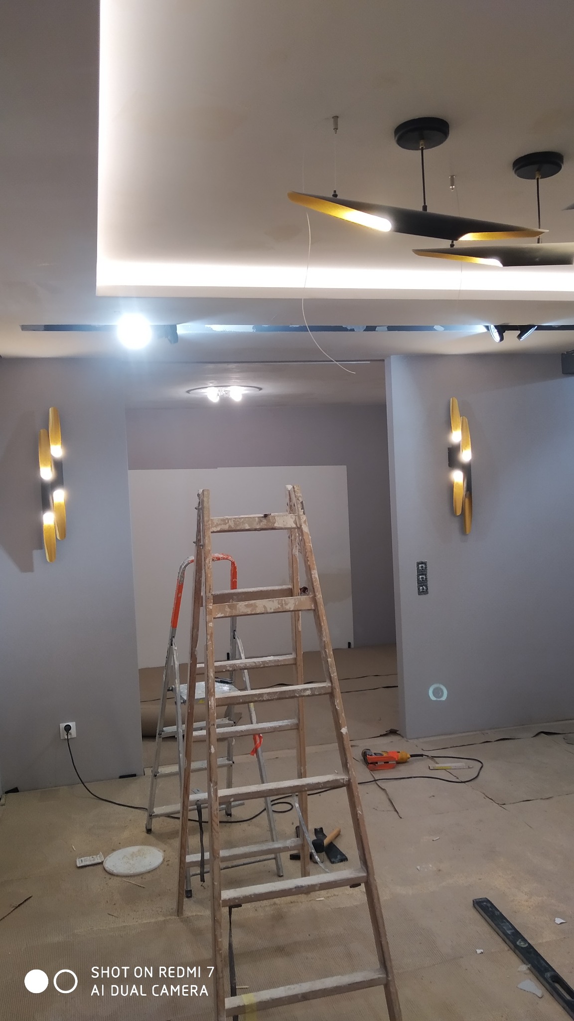 Wnętrze w trakcie prac wykończeniowych: dwie drabiny ustawione w pokoju z szarymi ścianami, nowoczesne złote kinkiety na ścianach, sufit z oświetleniem LED, widoczne narzędzia i elementy instalacji...