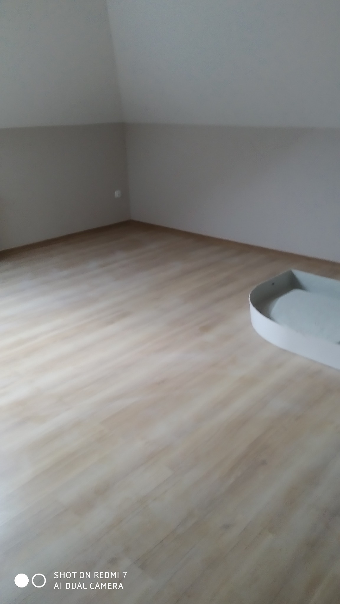 Pomalowane na szaro ściany i jasna podłoga imitująca drewno na poddaszu, z widoczną kuwetą malarską w rogu.