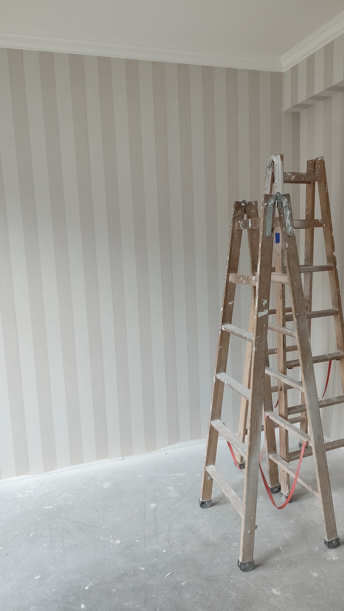 Ściana z tapetą w pionowe pasy w odcieniach beżu i bieli, wykończona listwą przysufitową, z dwiema drewnianymi drabinami malarskimi w tle, na podłodze widoczne ślady po remoncie.