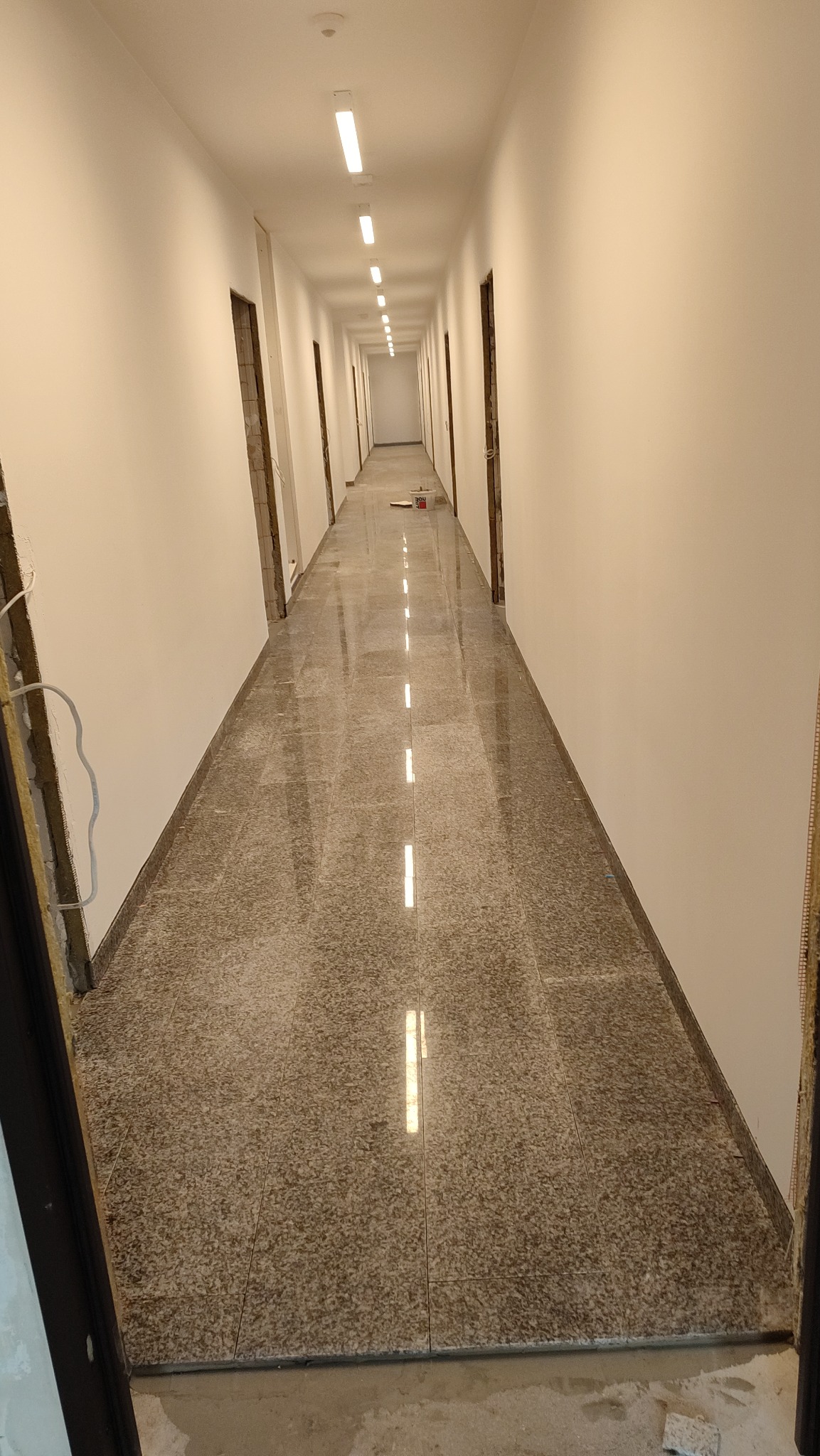 Długi korytarz w trakcie remontu, z nową, błyszczącą, brązowo-beżową podłogą z płytek imitujących kamień, świeżo pomalowanymi na biało ścianami i otworami drzwiowymi w stanie surowym.