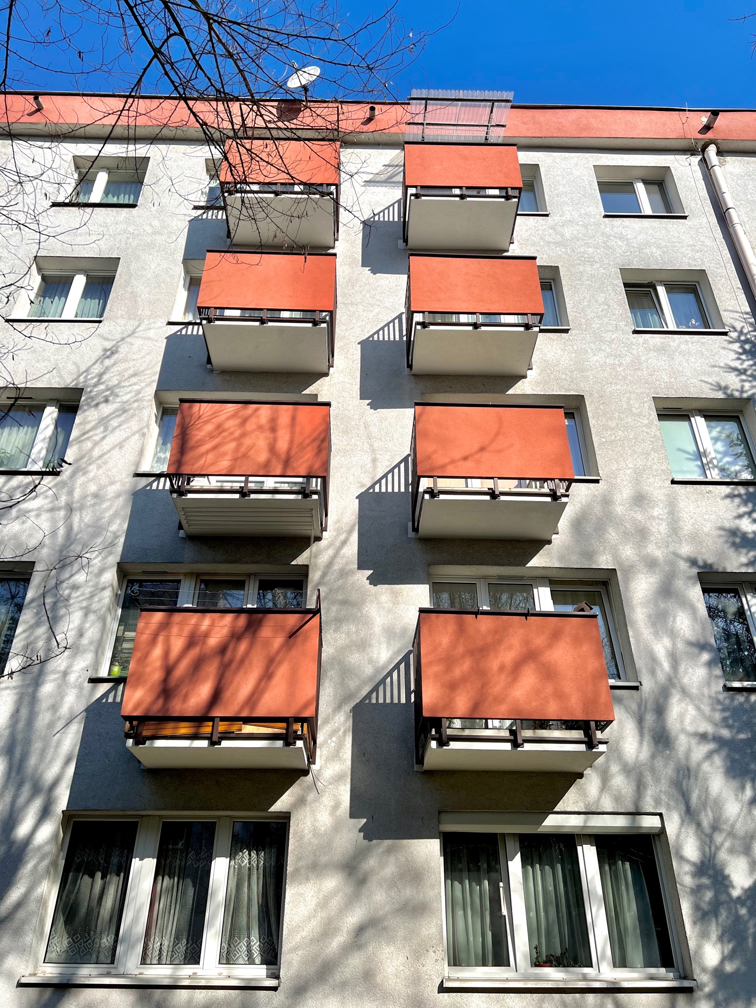 Fasada bloku z rzędem balkonów osłoniętych pomarańczowymi panelami, cień drzew na ścianie, błękitne niebo w tle.