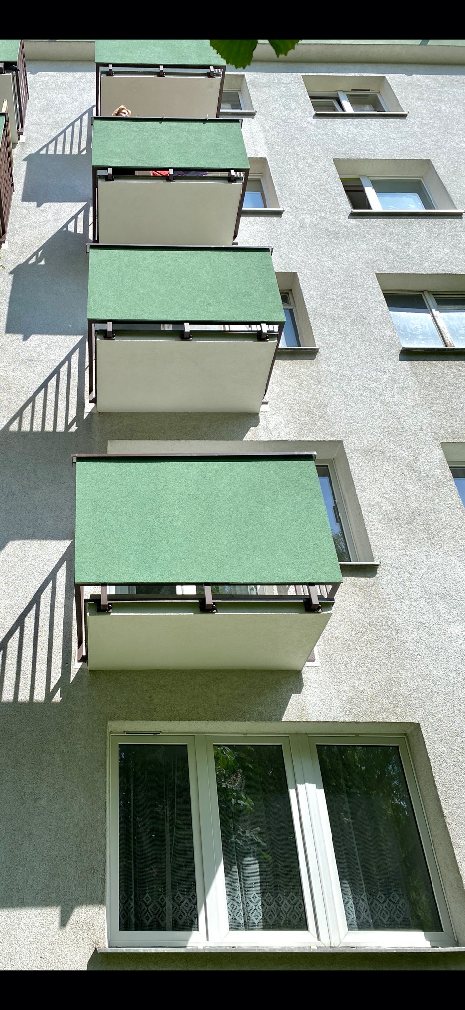 Fasada budynku z rzędem balkonów z zielonymi osłonami, cień balustrady na ścianie, okno z firanką na dole.
