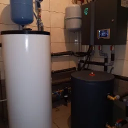 Instalacja z białym i czarnym zbiornikiem buforowym, szarym modułem sterującym z podświetlonym logo i wyświetlaczem, rurami i armaturą w kotłowni wyłożonej płytkami.