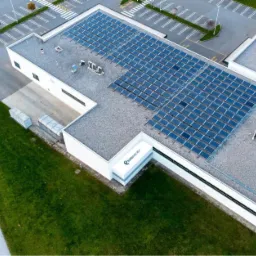 Widok z góry na budynek przemysłowy z zainstalowaną instalacją fotowoltaiczną na dachu, obok parking z samochodami, w tle drzewa.