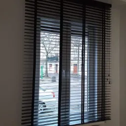 Okno z zamontowanymi czarnymi żaluzjami drewnianymi, widok na ulicę z samochodem i budynkami.