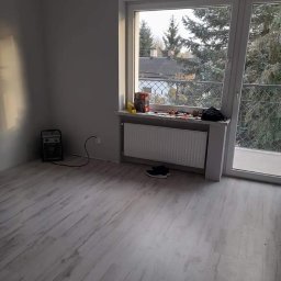 Flex Bud - Pokój po remoncie: świeżo pomalowane szare ściany, jasna podłoga imitująca drewno, grzejnik pod oknem z widokiem na drzewa, suszarka do butów w rogu.