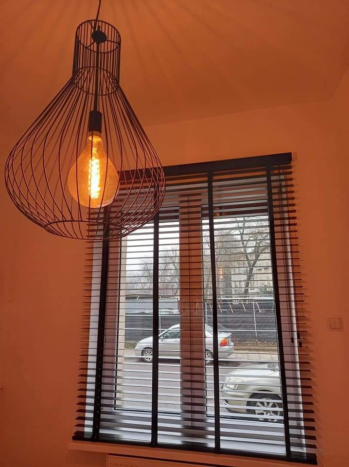 Nowoczesna lampa wisząca w kształcie klatki nad oknem z czarnymi żaluzjami poziomymi, widok na ulicę z dwoma samochodami, ciepłe oświetlenie wnętrza.