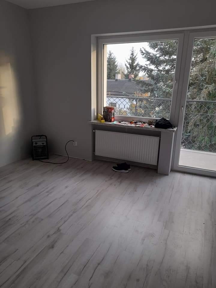 Pokój po remoncie: świeżo pomalowane szare ściany, jasna podłoga imitująca drewno, grzejnik pod oknem z widokiem na drzewa, suszarka do butów w rogu.