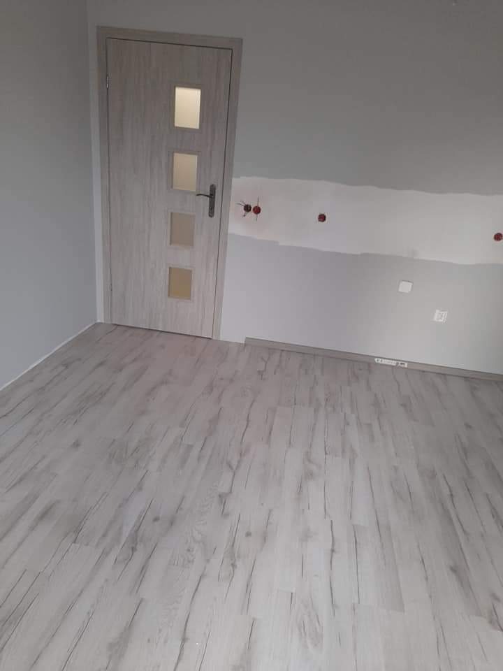 Wykończone wnętrze pokoju z jasną podłogą imitującą drewno, szarą ścianą z widocznymi punktami elektrycznymi i nową białą stolarką drzwiową z przeszkleniami.
