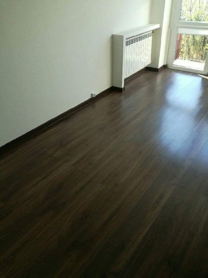 Wypolerowana, ciemnobrązowa podłoga z paneli, listwy przypodłogowe w kolorze podłogi, biały grzejnik pod oknem balkonowym w świeżo odnowionym pokoju.