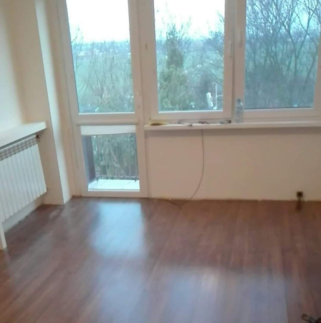 Pusty pokój z brązową podłogą, białymi ścianami i dużym oknem z widokiem na zewnątrz, grzejnik pod parapetem.