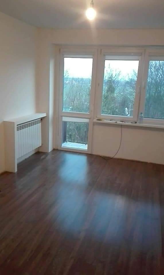 Pusty pokój po remoncie z ciemną podłogą, białymi ścianami i zabudowanym grzejnikiem pod oknem z widokiem na drzewa.