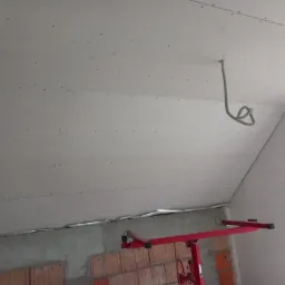 Montaż płyt kartonowo-gipsowych na poddaszu, widoczne łączenia płyt, wystający kabel elektryczny i podnośnik do płyt. Surowy stan wykończenia.