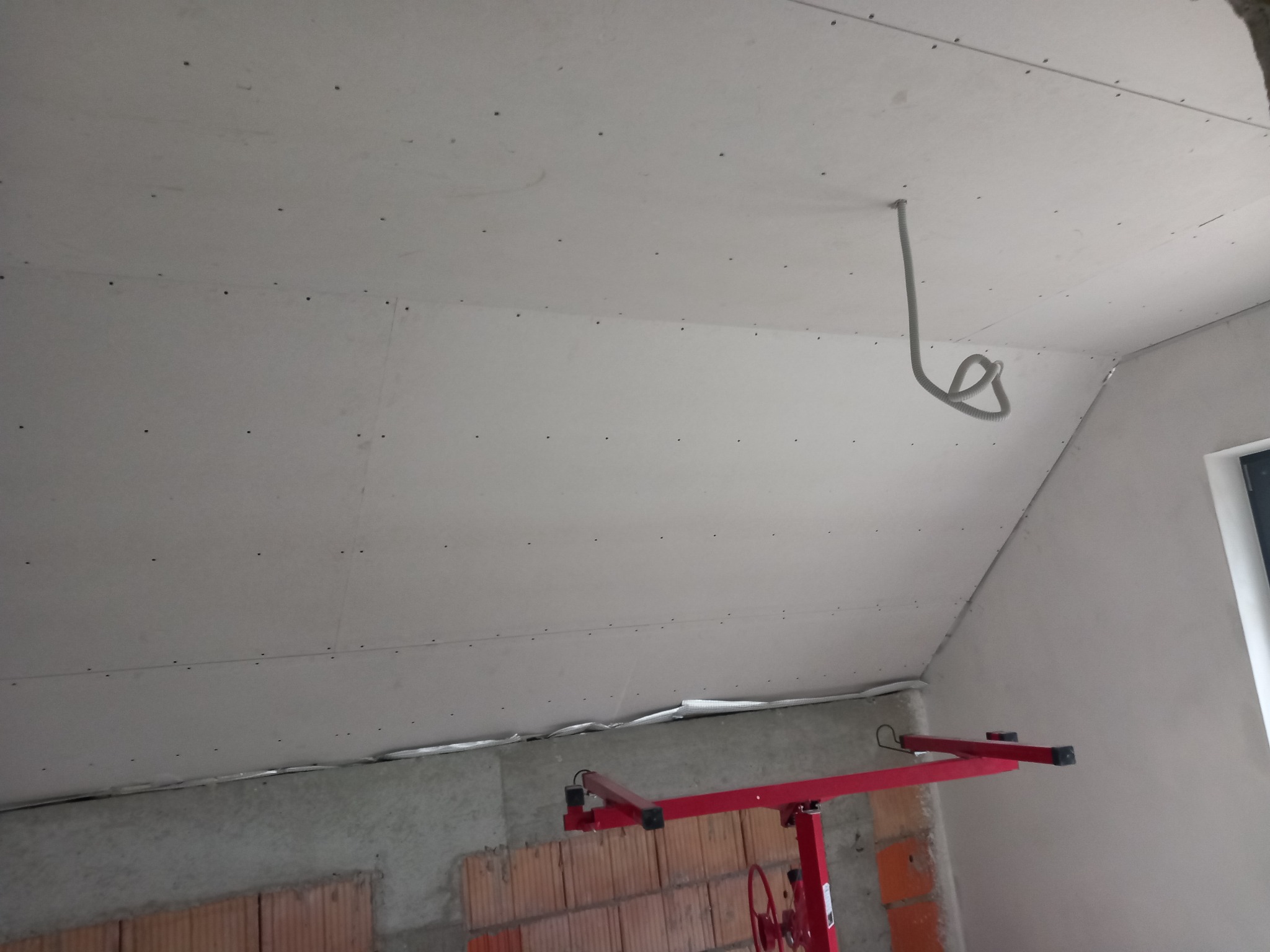 Montaż płyt kartonowo-gipsowych na poddaszu, widoczne łączenia płyt, wystający kabel elektryczny i podnośnik do płyt. Surowy stan wykończenia.