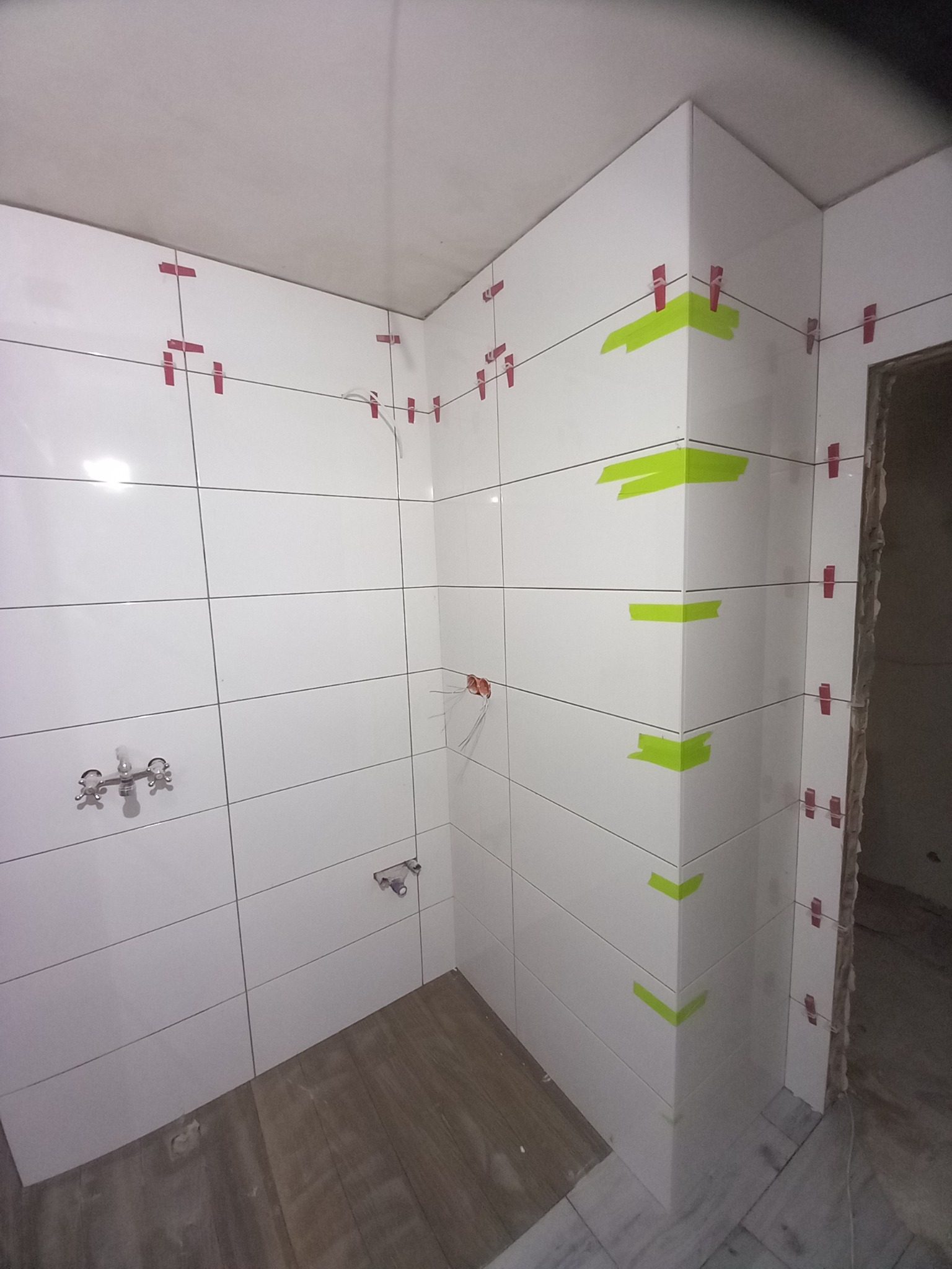 Białe, błyszczące płytki ceramiczne w trakcie układania w rogu łazienki, z użyciem czerwonych krzyżyków dystansowych i zielonej taśmy maskującej na krawędziach.