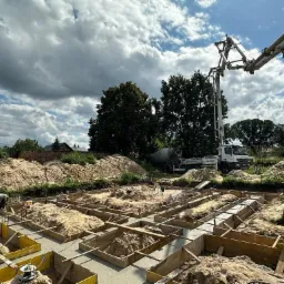 Wylewanie betonu w szalunkach fundamentowych pod budowę domu jednorodzinnego, widoczny betonowóz z pompą, pracownik w kasku oraz hałdy piasku.