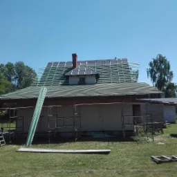 Dom jednorodzinny w trakcie montażu łat dachowych pod nowe pokrycie, widoczne rusztowania i materiały budowlane na trawniku.