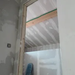 Wnętrze w trakcie wykańczania, widok przez surową ościeżnicę, ściany z płyt kartonowo-gipsowych, widoczny fragment instalacji elektrycznej, w tle dziecięce krzesełko.