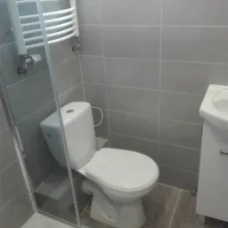 Szara łazienka po remoncie: widok na toaletę, umywalkę z szafką i grzejnik drabinkowy, szare płytki na ścianach i podłodze, kabina prysznicowa z przezroczystego szkła.