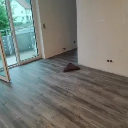 Nowe, szare panele podłogowe imitujące drewno ułożone w pustym pokoju z otwartym włazem rewizyjnym, widok na balkon i niedomalowane ściany.