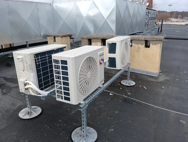Instalacja kilku zewnętrznych jednostek klimatyzacyjnych na dachu budynku, wspartych na metalowych konstrukcjach z okrągłymi podstawami, z widocznymi przewodami i większymi instalacjami w tle.