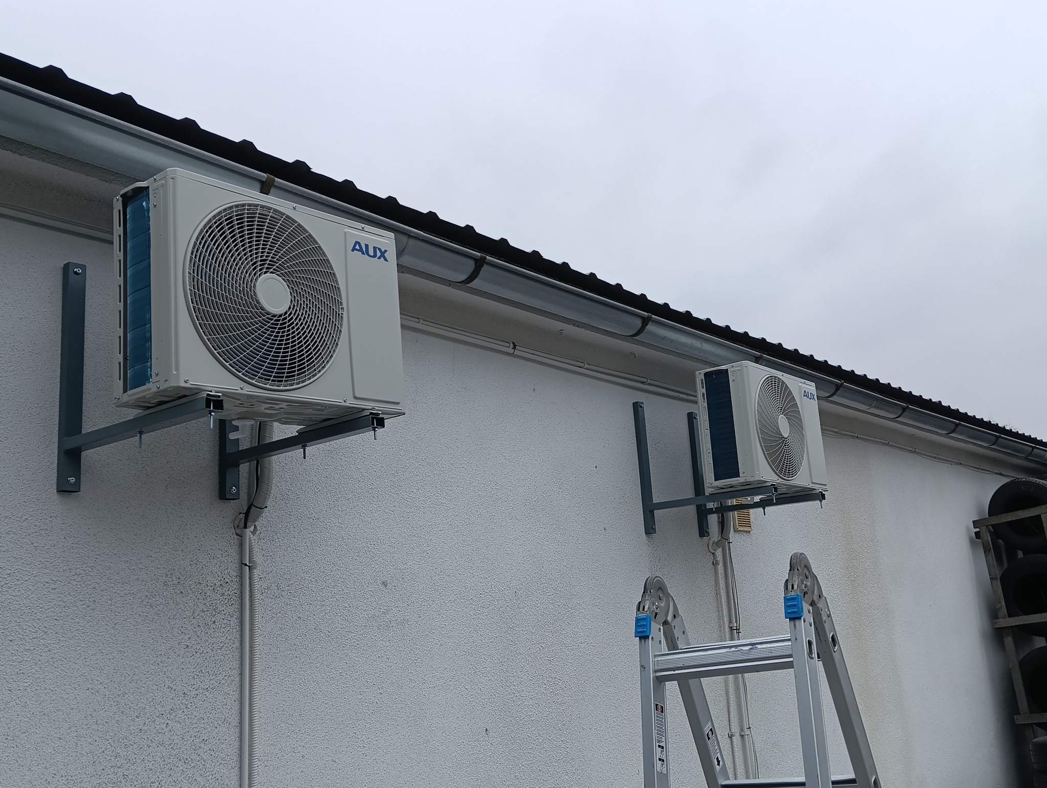 Dwa klimatyzatory marki AUX zamontowane na zewnątrz budynku z teksturą baranka, widoczna drabina oparta o ścianę.