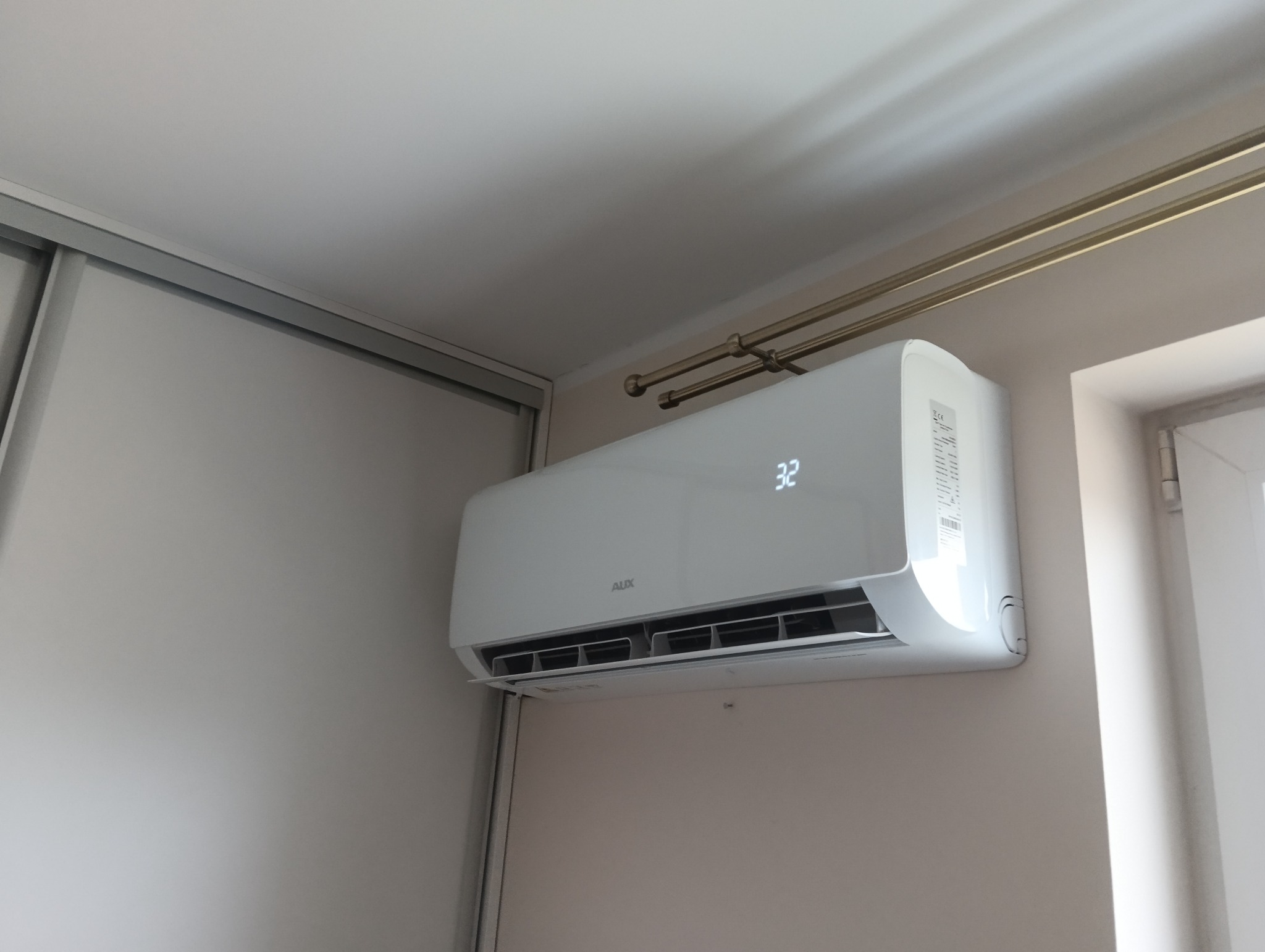 Biała klimatyzacja AUX zamontowana na ścianie w pomieszczeniu, widoczny wyświetlacz pokazujący temperaturę 32 stopnie Celsjusza, obok okno i metalowy karnisz.