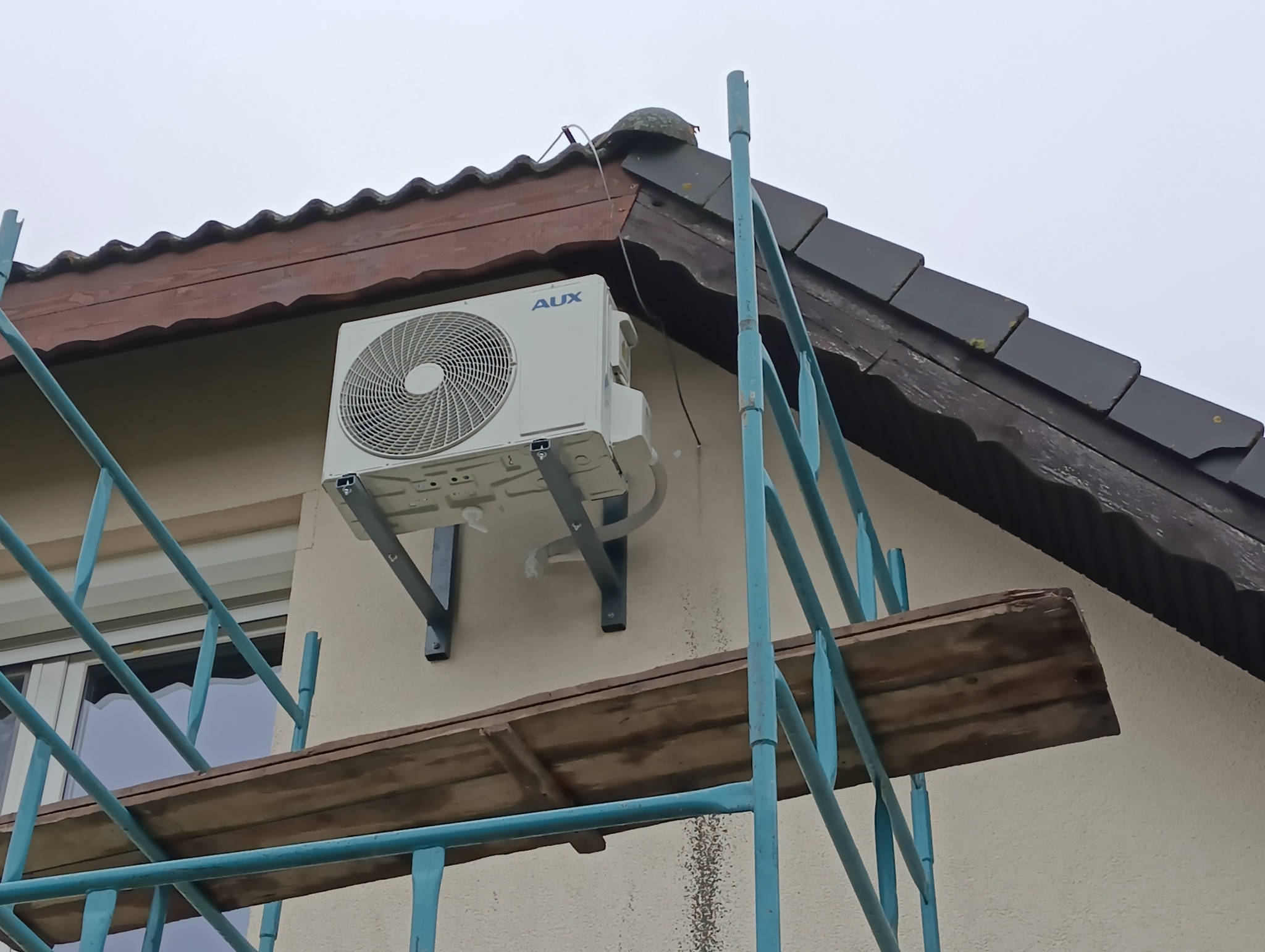 Zewnętrzna jednostka klimatyzacji AUX zamontowana na wspornikach na ścianie budynku, widoczna z rusztowania. Dach budynku z dachówką w tle.