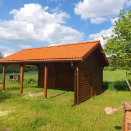 Drewniana konstrukcja z zadaszeniem z czerwonej dachówki, połączona z pergolą, na trawniku w słoneczny dzień; w tle widoczne drzewa i błękitne niebo z chmurami.