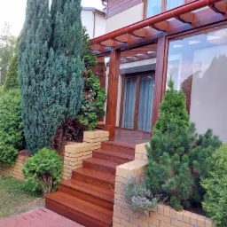 Wejście do domu z drewnianymi schodami, ceglanymi podmurówkami obsadzonymi zielenią oraz zadaszeniem z drewnianą konstrukcją i przezroczystym dachem.