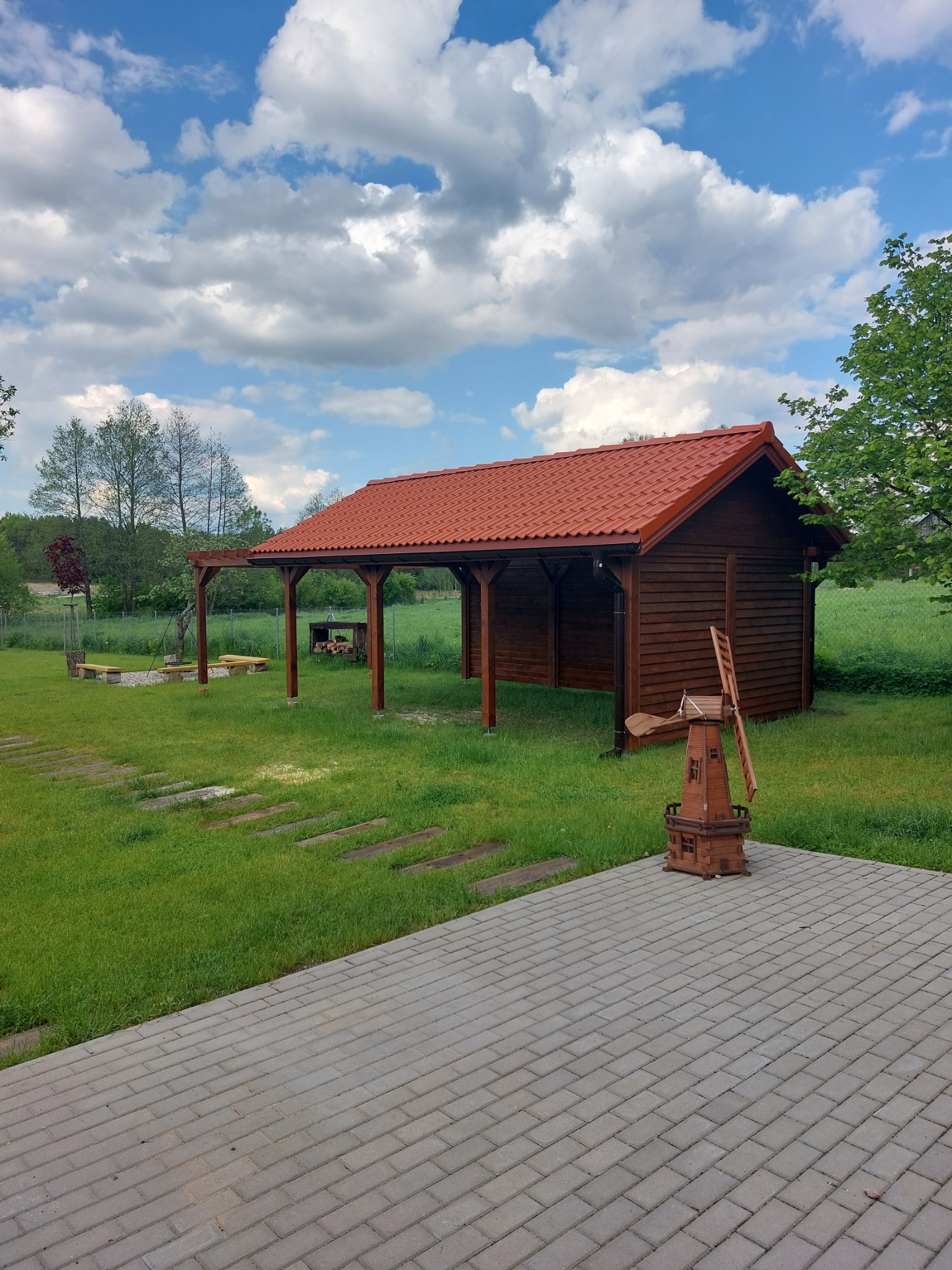 Drewniana altana z czerwonym dachem w ogrodzie, obok dekoracyjny wiatrak, widoczny fragment brukowanego tarasu i drewniane ścieżki.