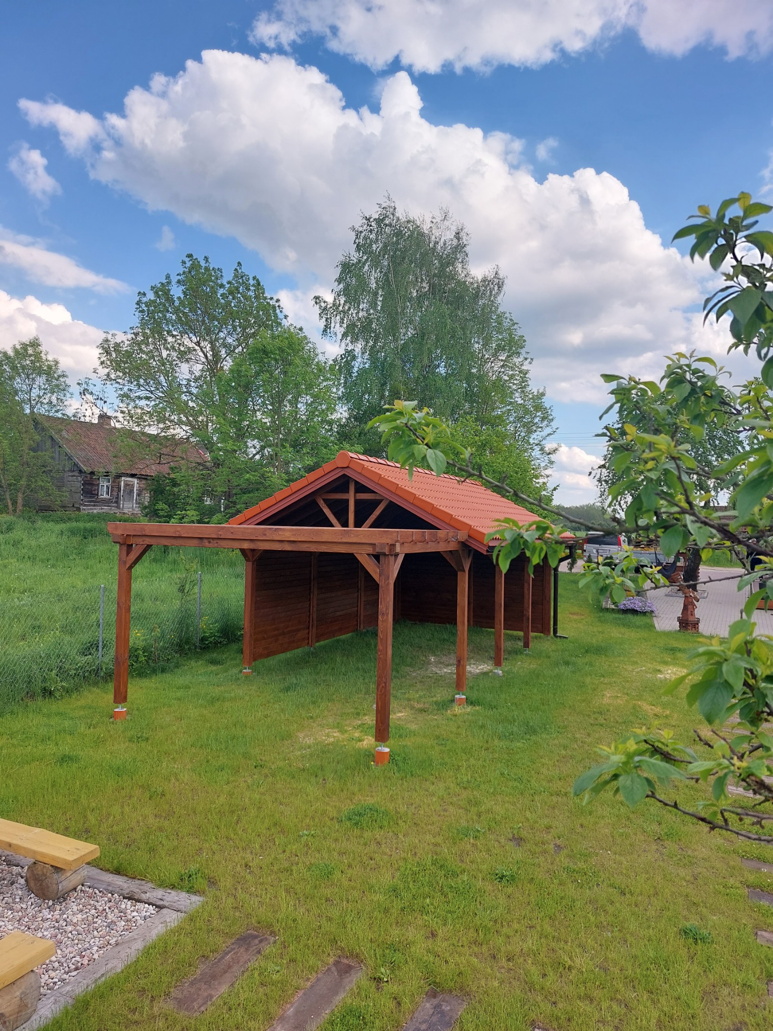Drewniana konstrukcja z czerwonym dachem, prawdopodobnie wiata lub zadaszenie tarasu, na zielonym trawniku w słoneczny dzień; w tle widoczny stary drewniany dom i błękitne niebo z chmurami.