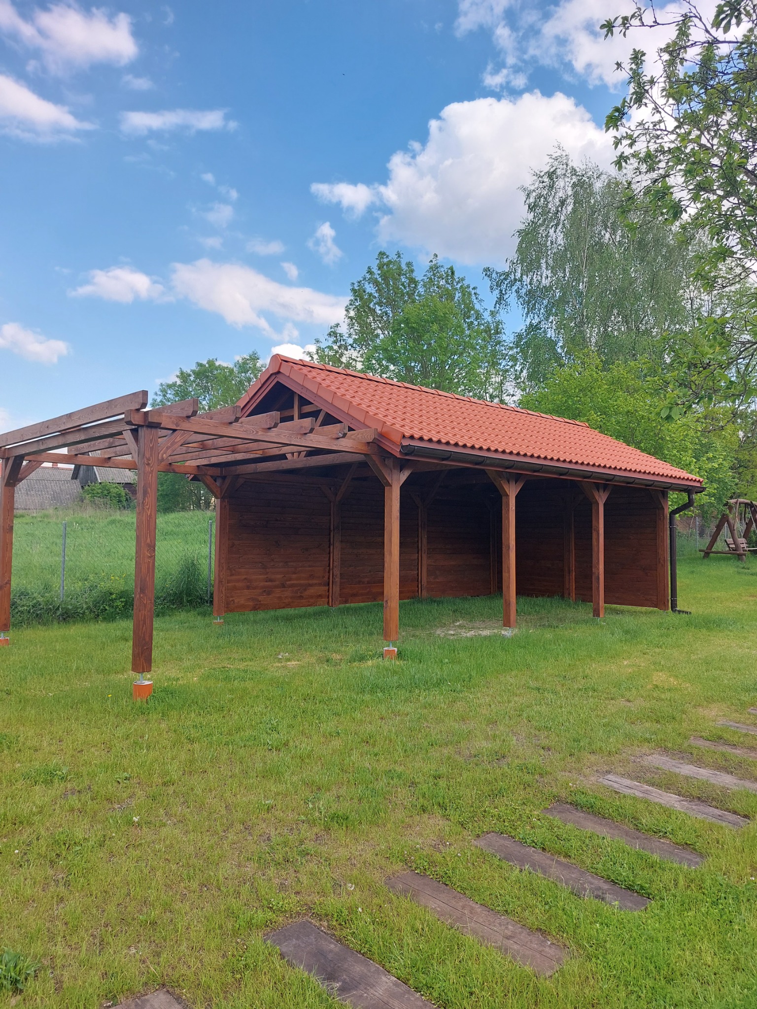 Drewniana konstrukcja z czerwoną dachówką i elementami pergoli w Wasilkowie, otoczona zielonym trawnikiem i drzewami.