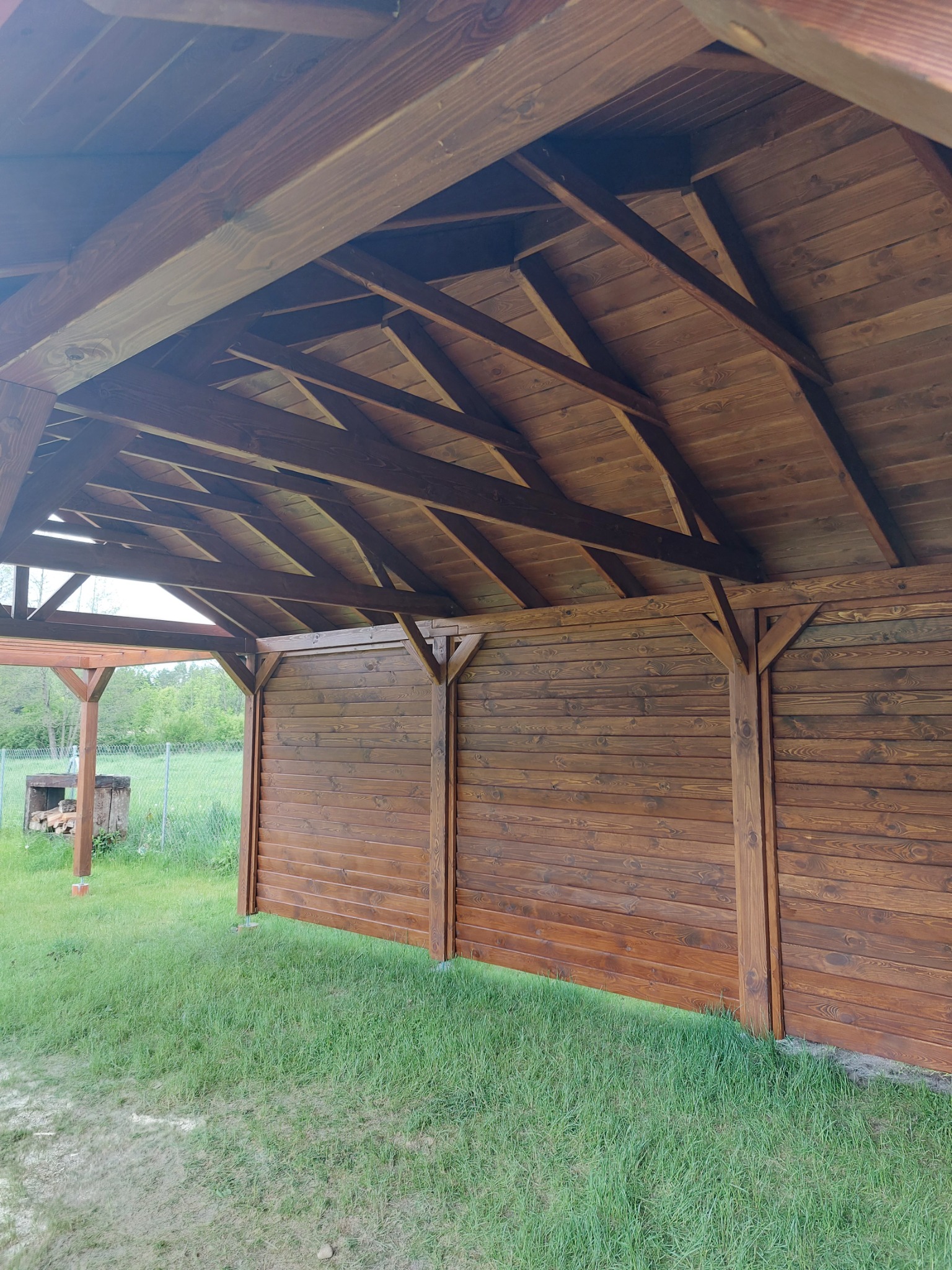 Wnętrze zadaszonej konstrukcji drewnianej, z widoczną konstrukcją dachu i ścianami z desek w kolorze ciemnego brązu, na zewnątrz zielony trawnik i częściowo widoczna pergola z paleniskiem.