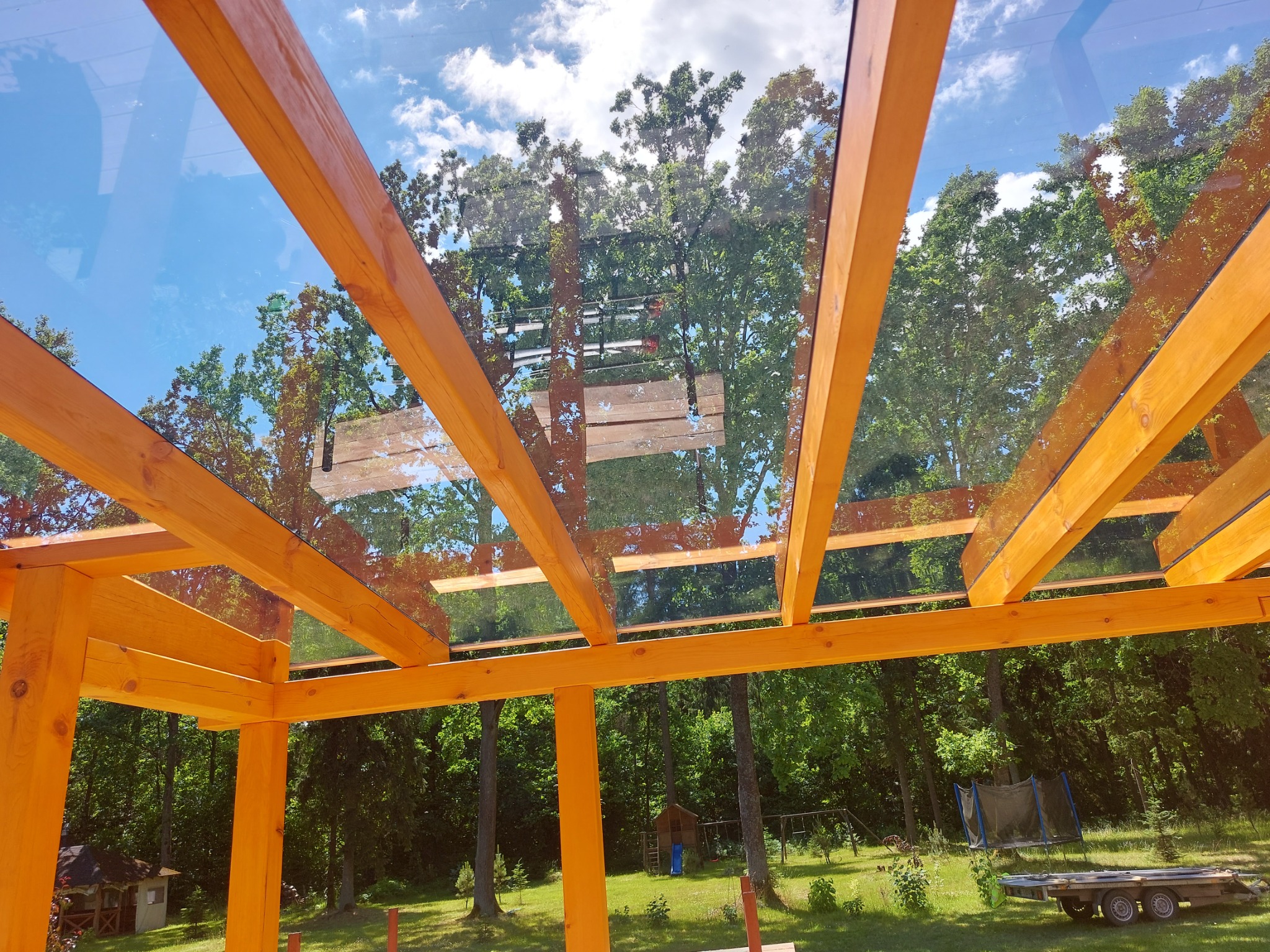 Widok z dołu na drewnianą konstrukcję z przeszklonym dachem, odbijającą błękitne niebo i korony drzew. W tle widoczny ogród z domkiem dla dzieci i trampoliną.