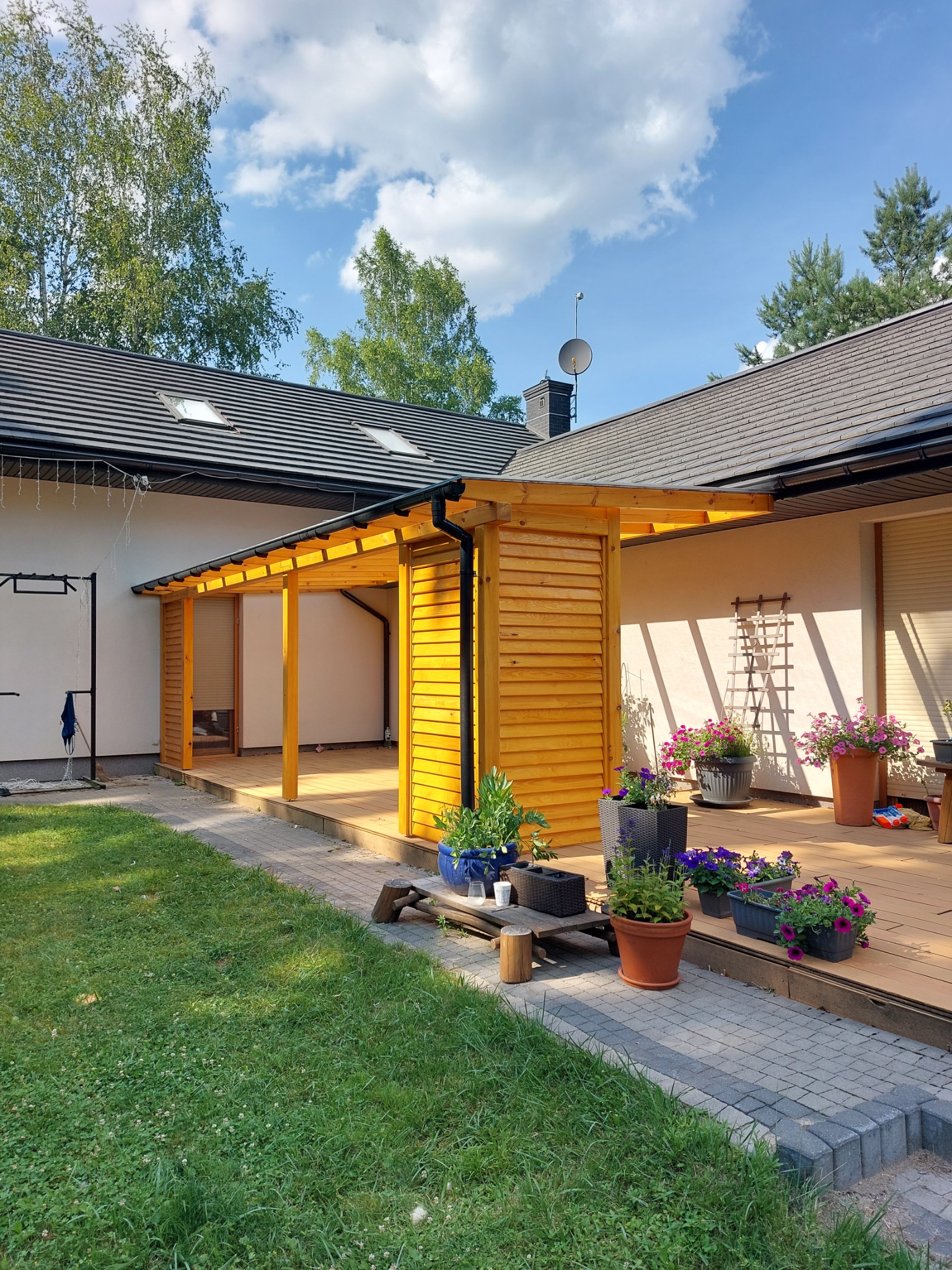 Drewniana konstrukcja przylegająca do domu, prawdopodobnie zadaszenie tarasu lub weranda, z widocznymi elementami boazerii i drewnianej podłogi, otoczona zielonym trawnikiem i kwiatami w donicach.