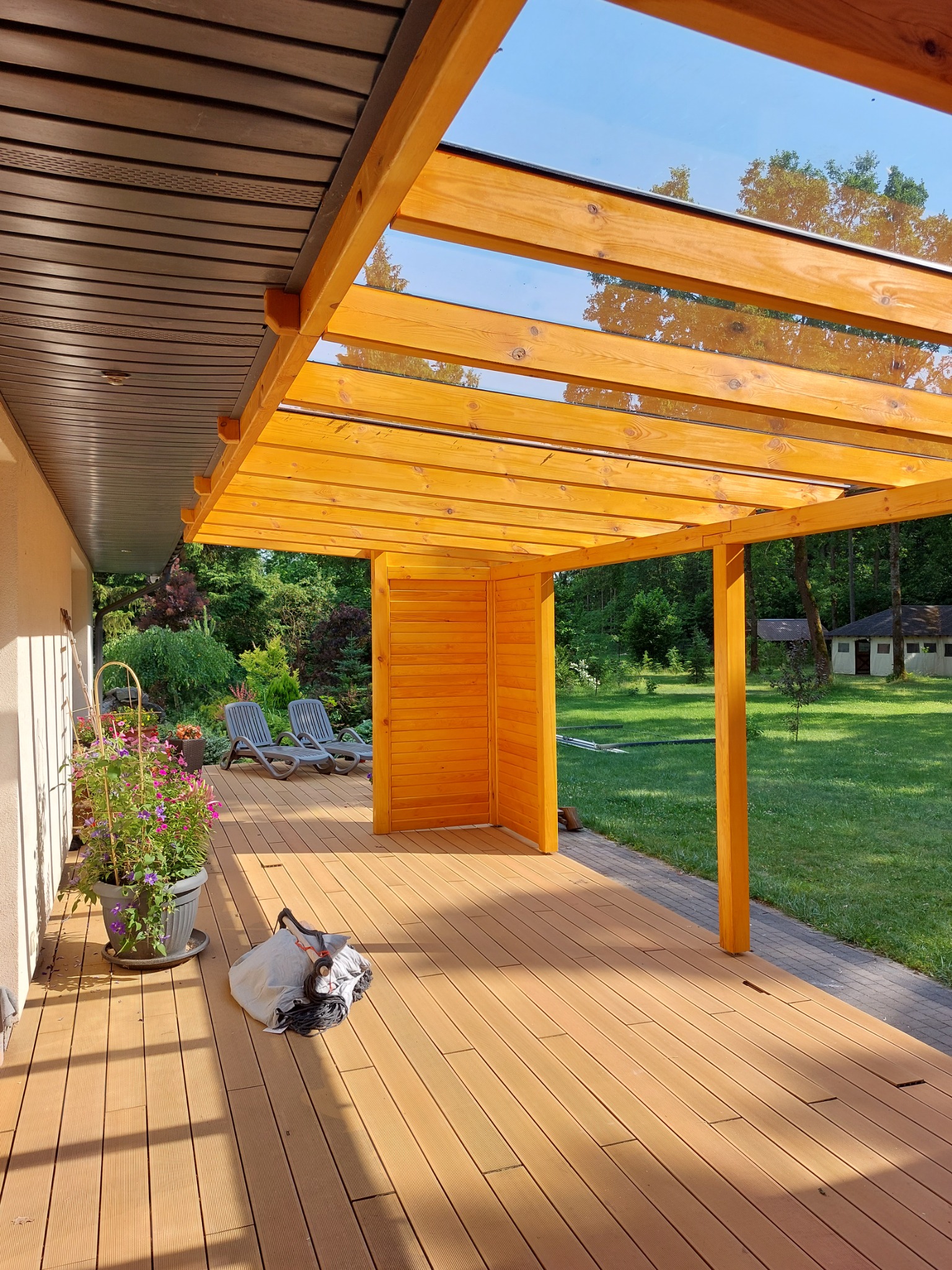 Zadaszony taras z drewnianą konstrukcją i przezroczystym dachem, z widokiem na ogród, leżaki i kwiaty w donicy, z torbą leżącą na podłodze.