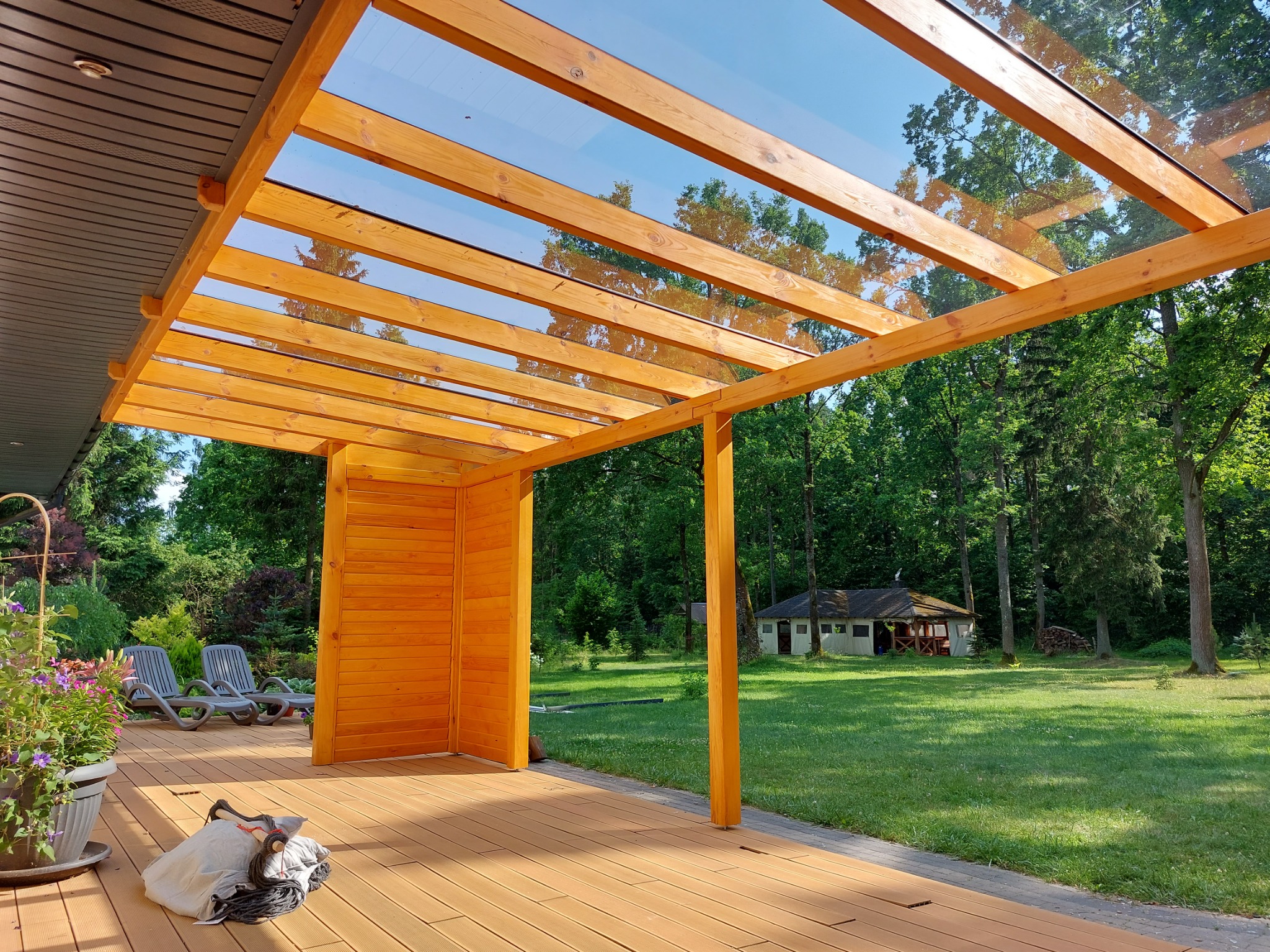 Zadaszony taras z drewnianą konstrukcją i szklanym dachem, widok na zielony ogród i domek w tle.
