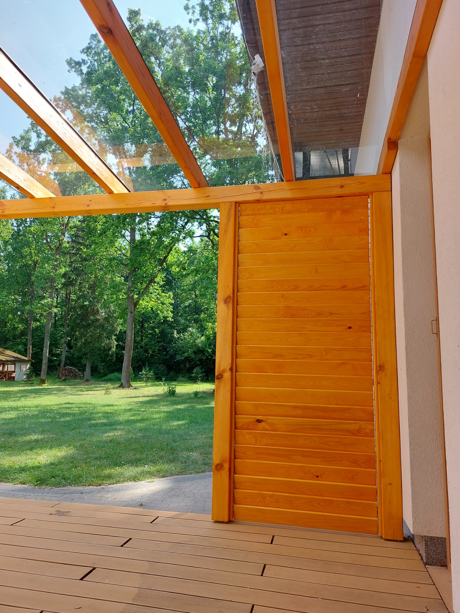 Drewniana pergola z przeszklonym dachem i przesuwnymi drewnianymi panelami, widok na zielony ogród z domkiem letniskowym w tle.