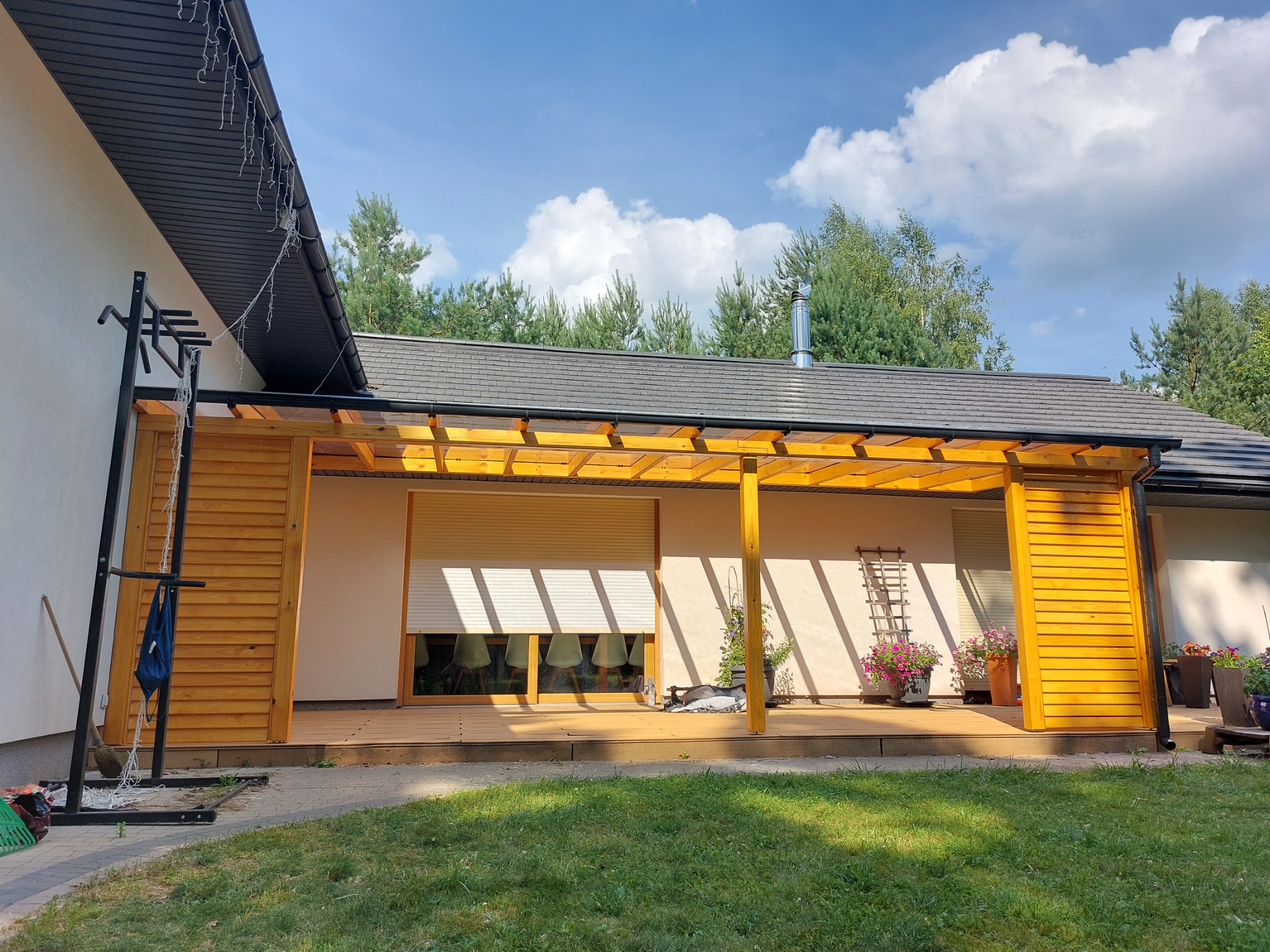 Zadaszony taras z drewnianą konstrukcją, przesłonięty częściowo drewnianymi żaluzjami, widok na ogród, okno z roletą i krzesłami w tle.