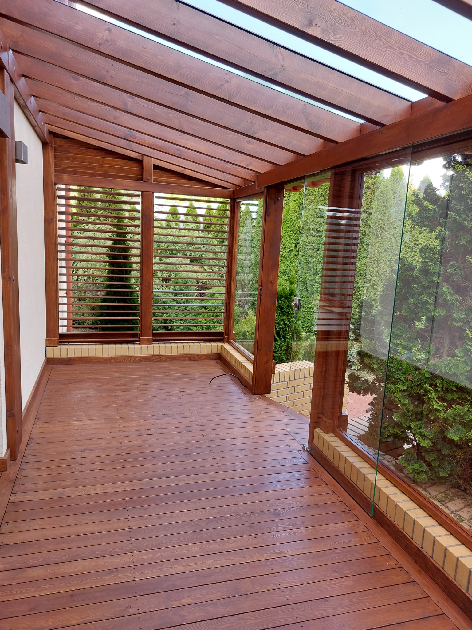 Zadaszony taras z drewnianą podłogą i konstrukcją, częściowo osłonięty szklanymi panelami i drewnianymi żaluzjami, widok na zieleń z zewnątrz.