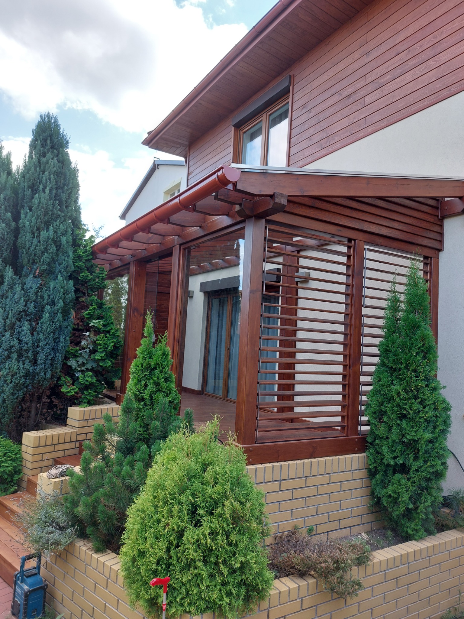 Drewniana konstrukcja z lamelami przy domu, z brązowymi elementami, otoczona zielenią iglaków i ceglanym murkiem, widok z zewnątrz.