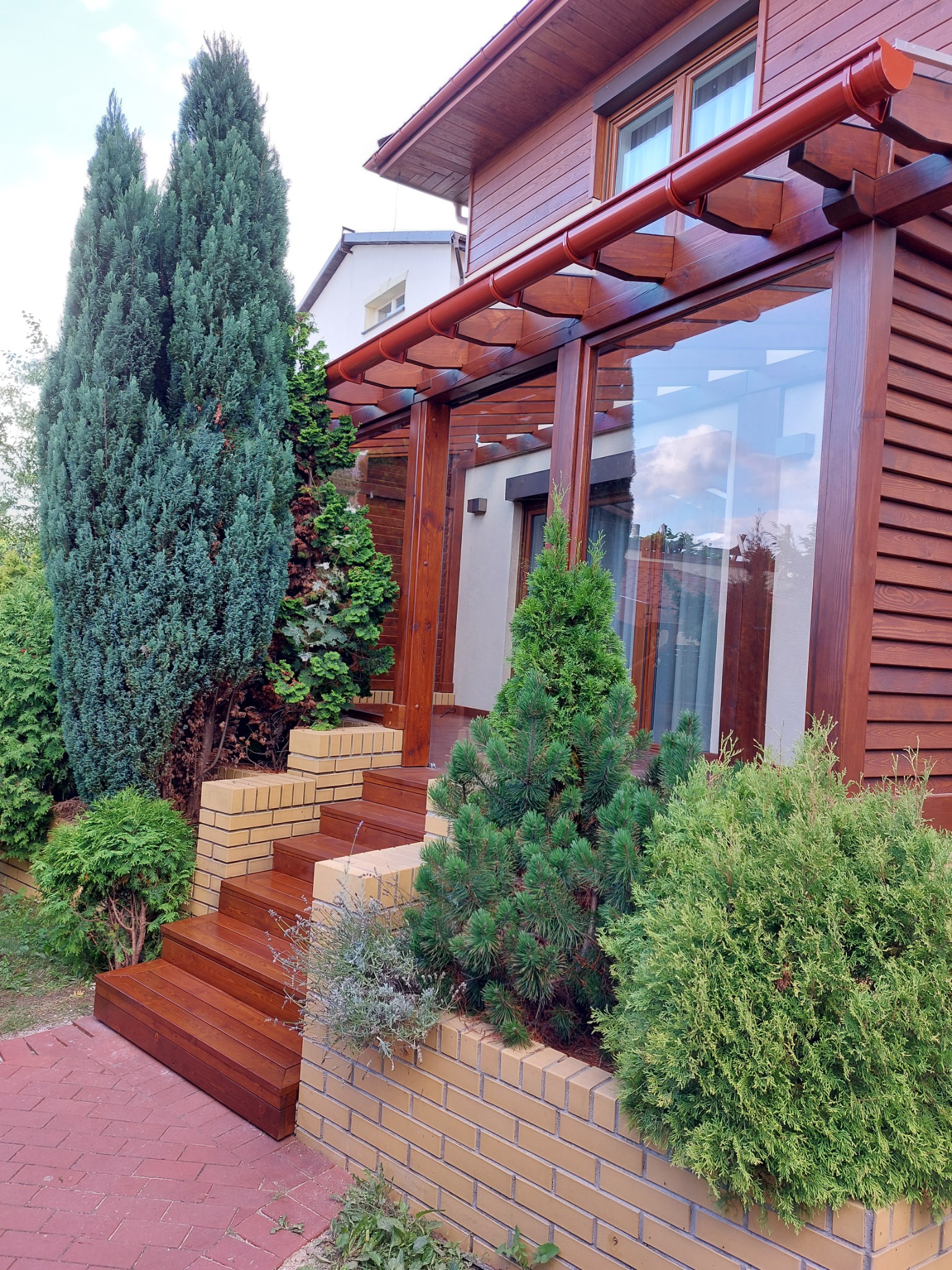 Wejście do domu z drewnianą elewacją, schodami z ceglanymi obrzeżami i zadaszeniem z przezroczystym dachem, otoczone różnorodną zielenią, w tym wysokimi cyprysami.