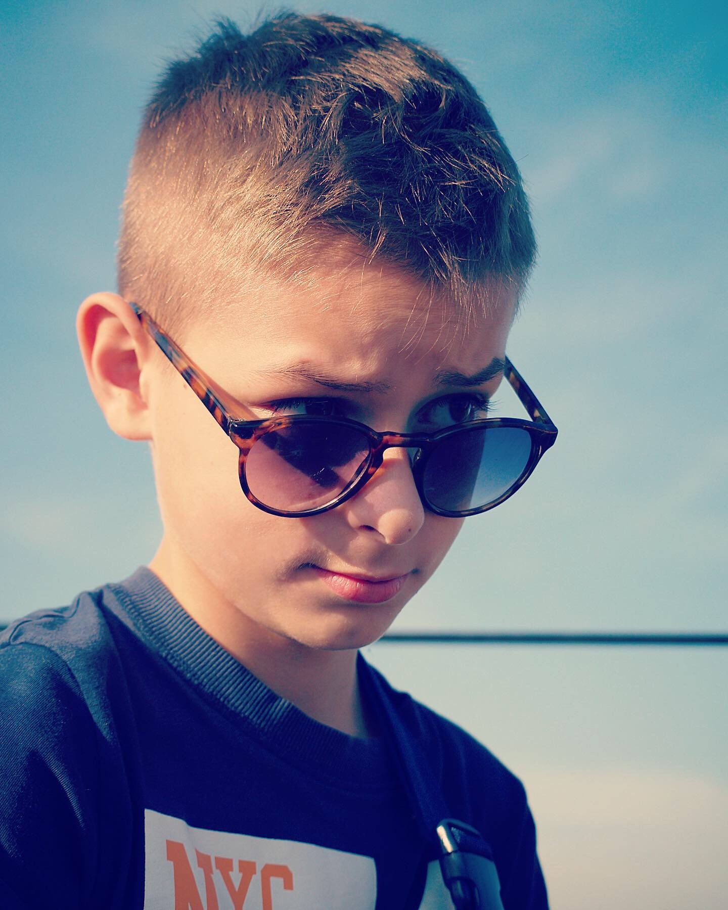 Portret chłopca w okularach przeciwsłonecznych z wzrokiem skierowanym w bok, na tle błękitnego nieba.