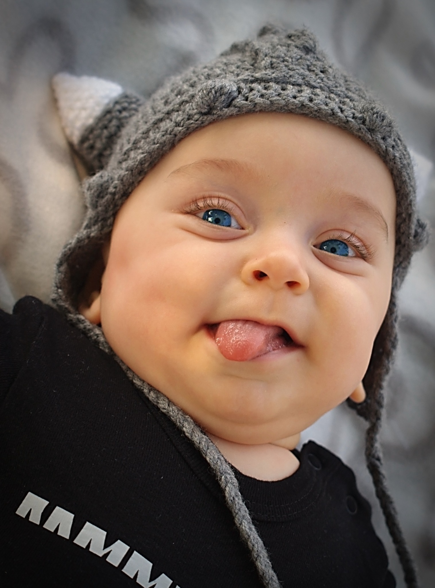 Uśmiechnięty niemowlak z niebieskimi oczami w szarym, dzierganym kapeluszu z uszkami, wystawia język.