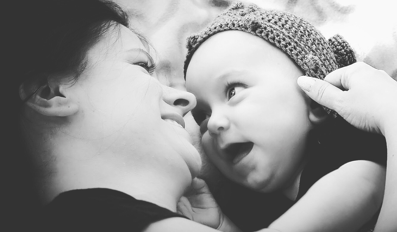 Czarno-białe zbliżenie matki z długimi rzęsami i uśmiechem patrzącej na niemowlę w dzierganej czapce, którego ręka dotyka.