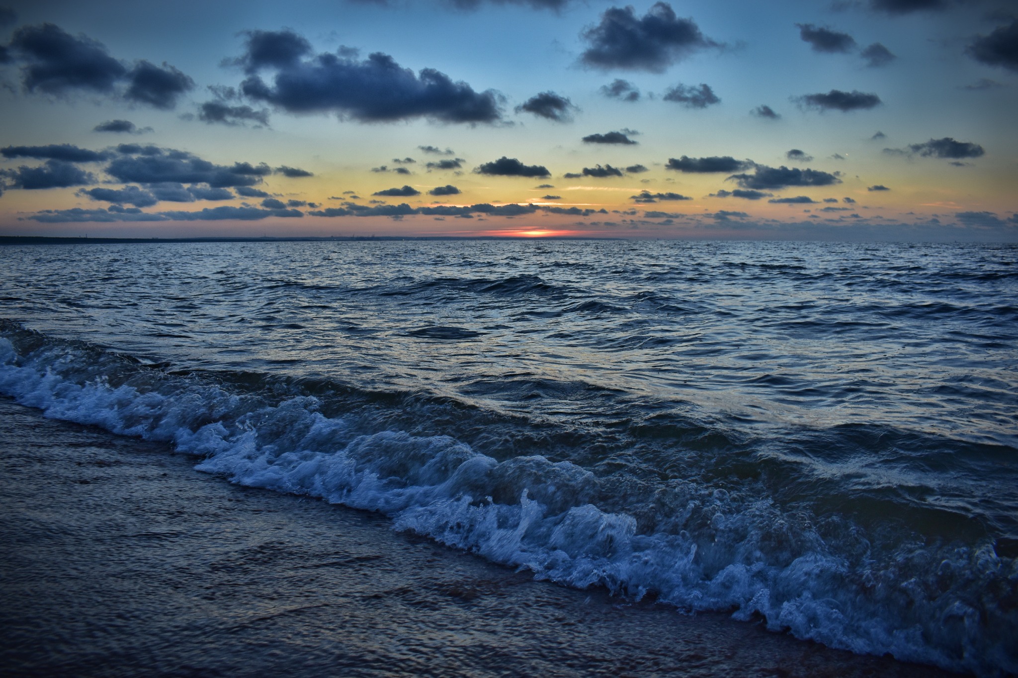 Spokojne morze o zmierzchu, z delikatnymi falami rozbijającymi się o brzeg, ciemne chmury na niebie i pas ciepłego światła zachodzącego słońca na horyzoncie.