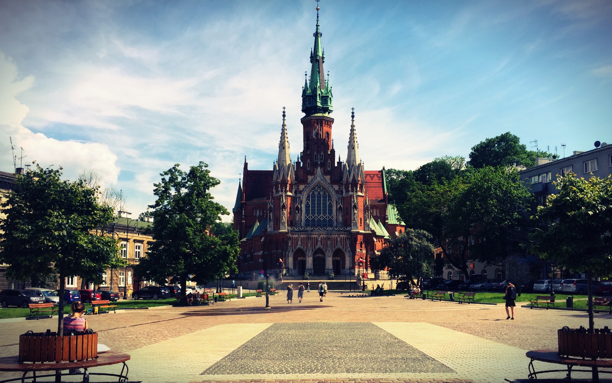 Bazylika św. Józefa w Krakowie z perspektywy placu, widoczne osoby spacerujące i odpoczywające na ławkach, drzewa i budynki w tle, błękitne niebo z chmurami.